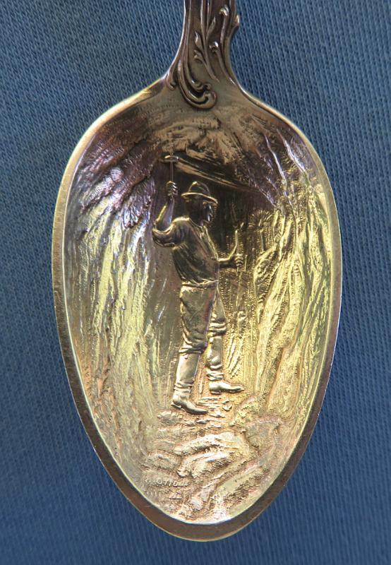 Souvenir Mining Spoon Prescott AZ Bowl.JPG - SOUVENIR MINING SPOON PRESCOTT AZ - Sterling silver souvenir spoon, features handle with miner and gold rocker box at top above picks and ore bucket, bowl embossed in deep relief with miner single jacking with artist marking H. OTTO in small letters near tip, back of handle marked PRESCOTT,ARIZONA at top above figure of cowboy on horse, back marked STERLING with makers mark of George E. Homer (Boston, MA 1875 - ?) and COPYRIGHTED, measures 5 1/2 in. length , weighs 37.3 gr. [Prescott is located in the Bradshaw Mountains of central Arizona, approximately 100 miles north of Phoenix.  The new town of Prescott was named in honor of historian William H. Prescott during a public meeting in 1864 and was officially incorporated in 1881. Prescott twice served as the capital of the Arizona Territory, once till November 1867 and again from 1877 to 1889 when the capital was moved to Phoenix.   Gold was discovered in creeks in the Agua Fria River Basin near Prescott in 1863.  Joseph Walker and a party of prospectors had set out from the California gold fields, possibly fleeing from conscription in the Confederate or the Union Army during the Civil War.  They found gold placers and, shortly afterward, lode deposits.  These discoveries were in what collectively became the Big Bug Mining District a few miles southeast of Prescott.  When the railroad arrived around 1898 transportation of equipment, minerals, and people became easier and cheaper.  Gold, silver, lead, and/or copper were produced from several famous mines including the Poland, McCabe, Silver Belt, Little Jessie, Henrietta/Big Bug, Blue Bell, Boggs/Iron Queen, and Iron King.  Several smelters were also built in the area.  The World War I era saw the greatest production from the district; however, the post-war drop in metal prices caused many mines to close.  The Iron King was the last of the major mines in the district.  After a series of different owners, it was operated by the Shattuck-Denn Mining Company from 1942 until it closed in 1969.  Prospectors staked claims in the Jerome District approximately 35 miles northeast of Prescott in 1876.  In 1882 the United Verde Copper Company was formed and mining started.  The rich oxidized ores produced copper, gold, and silver.  Transportation costs were very high until William A. Clark of Butte, Montana fame bought the company and brought the railroad to Jerome.  The United Verde Mine prospered and became the largest copper mine in the territory.  The original smelter built on unstable ground adjacent to the mine was replaced by a larger, more efficient one in Clarkdale.   In 1912, the Little Daisy Mine near the United Verde was purchased by James S. Douglas.  In 1914 and 1916 rich ore bodies were discovered.  A smelter was built in Clemenceau near the current town of Cottonwood.  The United Verde Extension Mining Company mined out the extension in 1930.  Phelps-Dodge purchased the United Verde and operated an open pit until mining ceased in 1953.  Today, the Douglas Mansion adjacent to the Little Daisy Shaft is part of the Jerome State Historic Park.  Jerome was designated a National Historic District in 1967.  Many of the historic buildings in Jerome have been converted to shops and eateries catering to tourists.  The Jerome Historical Society Museum occupies one of the buildings.  The Bagdad Mining District is located about 40 miles west of Prescott.  It has a rich history of mining including such famous mines as the Old Dick, Copper King, Copper Queen, and the Hillside among others.  The Bagdad deposit was discovered in 1862 and the claims were patented in 1889.  It was owned by a series of companies.  Exploration drilling was started in 1919 and the first mill was constructed in 1928.  During World War II, a 2000 ton per day mill was constructed with funds from the Reconstruction Finance Corporation. In the late 1940’s, the underground mine using block caving was converted to an open pit.  The town of Bagdad was a company town.  In 1973 the Bagdad Copper Corp. merged with the Cyprus Mines Corp. to form the Cyprus Bagdad Copper Company.  It later became part of Phelps-Dodge.  As a result of the 2007 merger, the mine is now operated by Freeport-McMoRan Copper and Gold Corporation. ] 